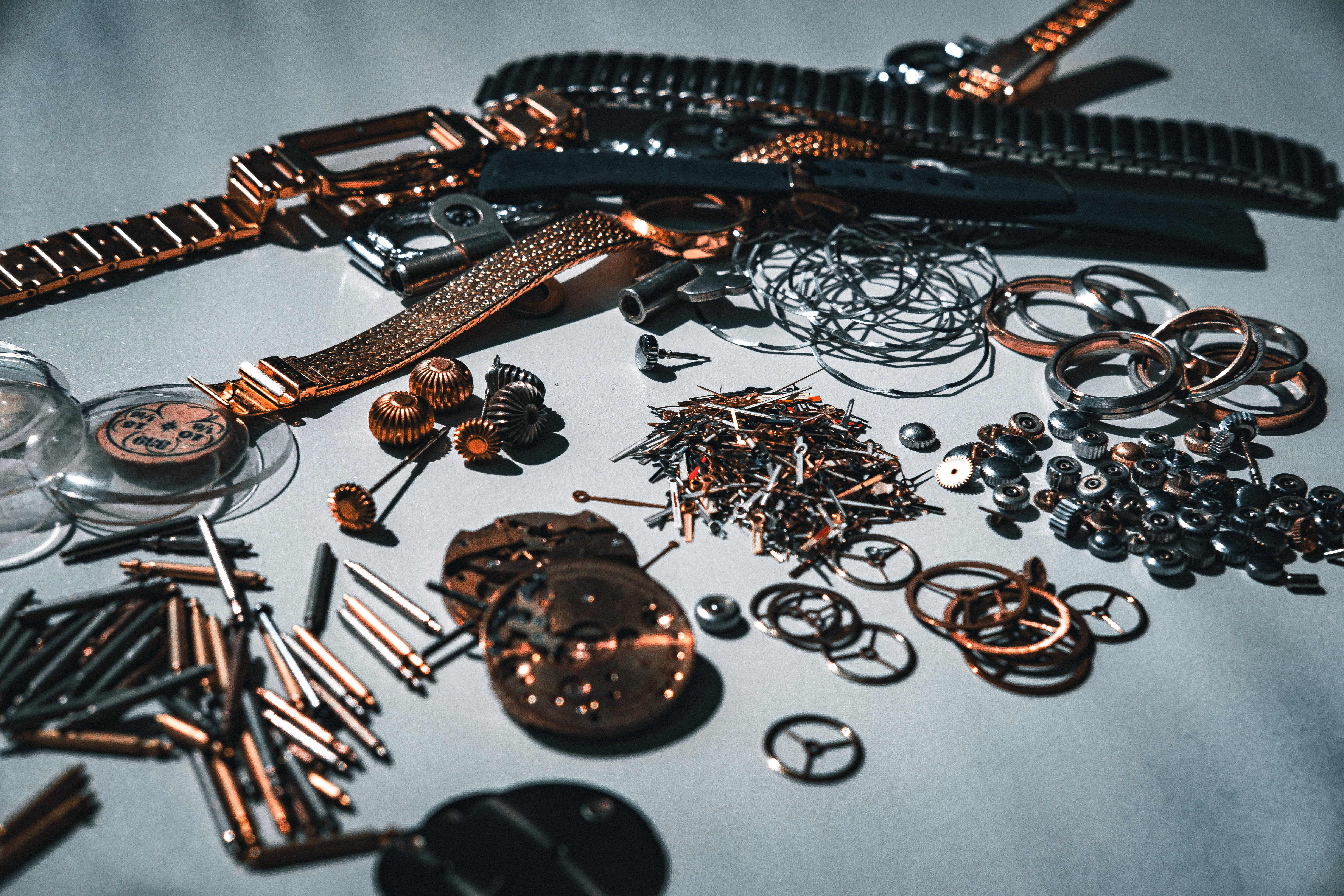 Old, metal watch parts roughly organized into small heaps by type. There are crowns, balance wheels, hands, movements, bracelets, pins, glass covers, circular springs in bunches, one type of part mixing with the next while still forming distinct clusters.