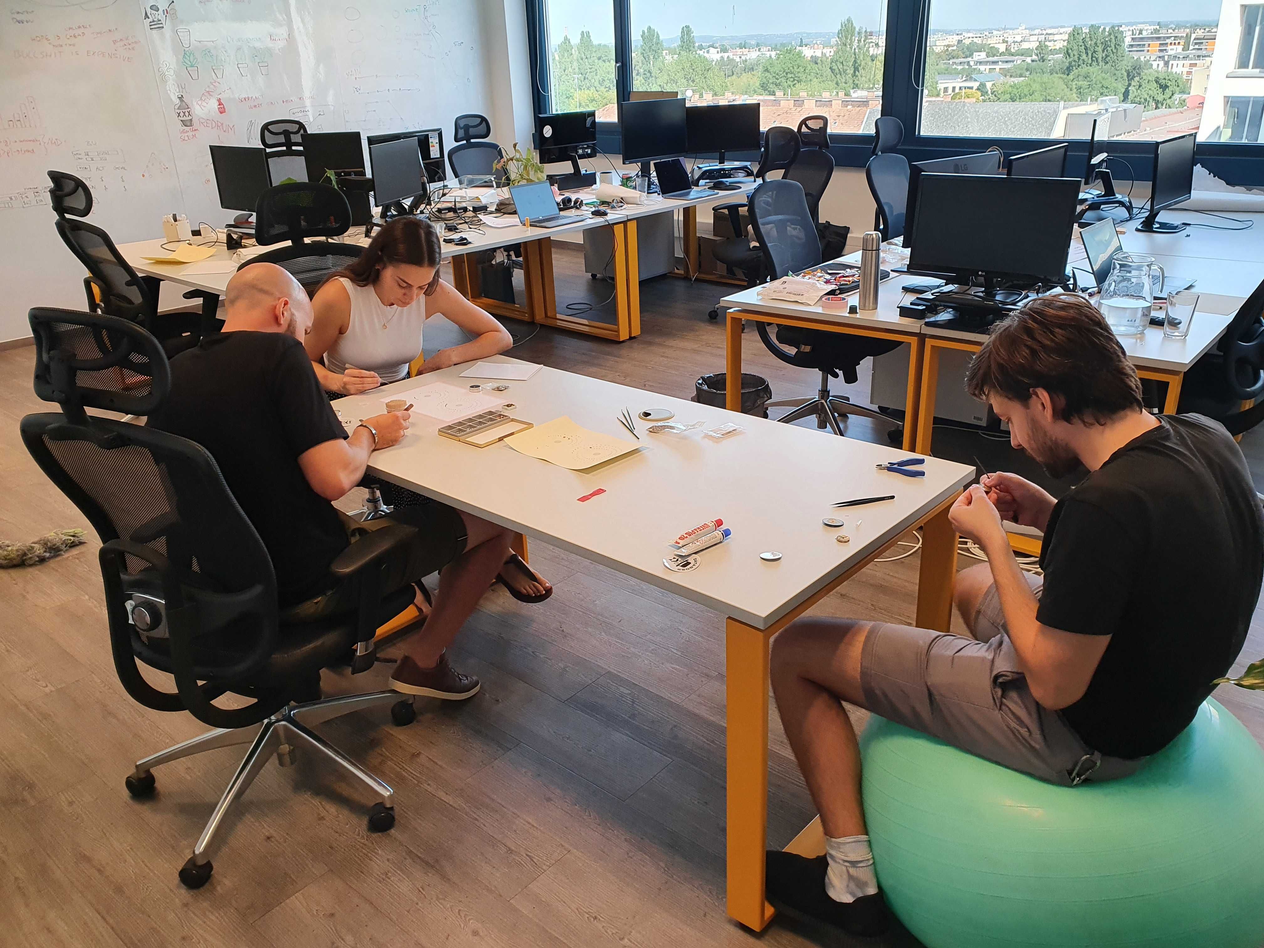Wide shot of the three authors working on the piece around a table in an office setting. All of them are slightly hunched, focused on the tiny parts they are gluing together and onto the paper. The other chairs and workstations in the office space are empty.