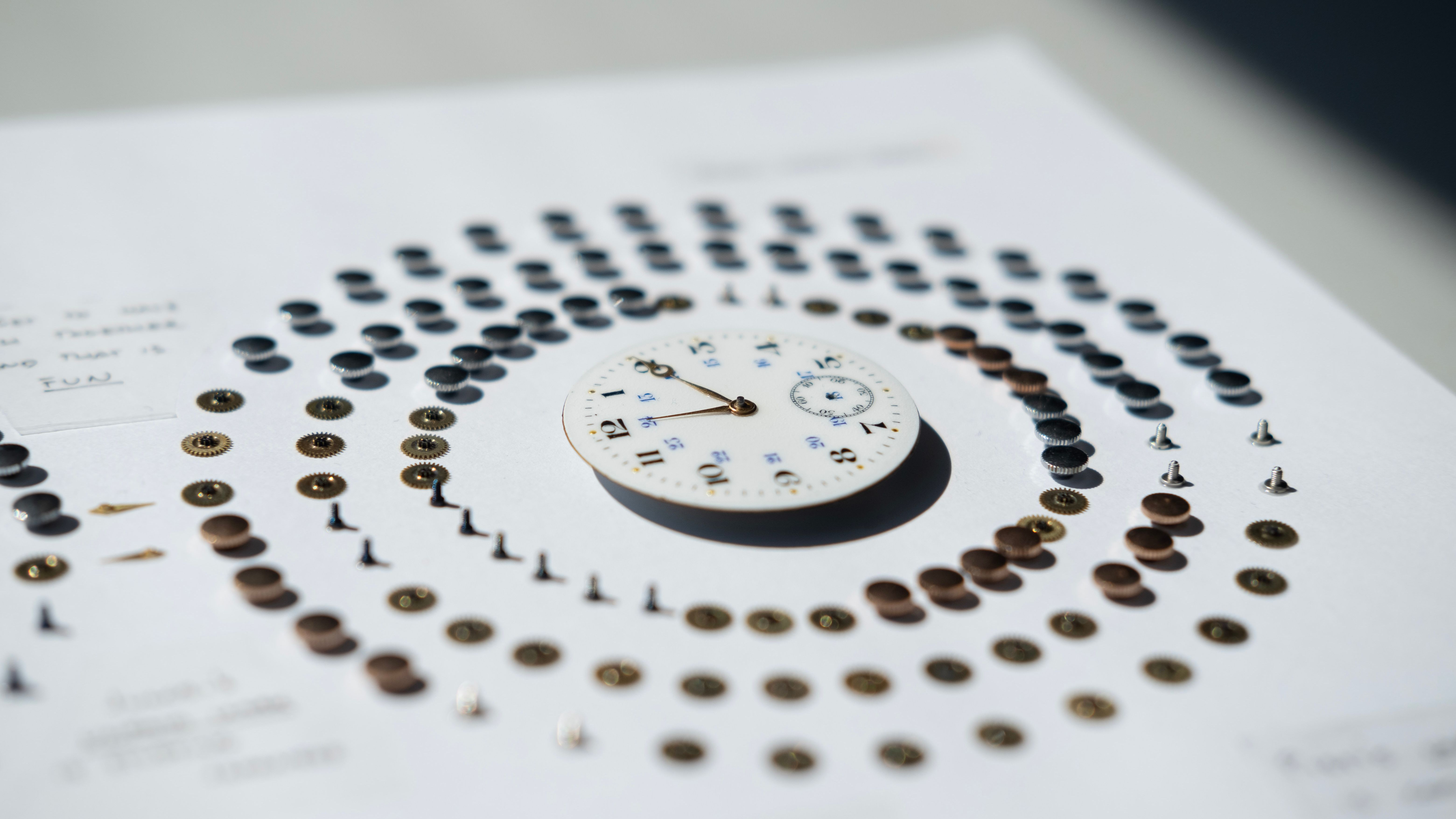 Closeup of the finished work, showing the dial around which the afternoon is visualized. The watch face is white and has a modern look with bold black and tiny blue numbering. At the part denoting 12 PM to 1 PM there are 9 brass gears next to each other showing that the authors have fun in their lunchtime together.